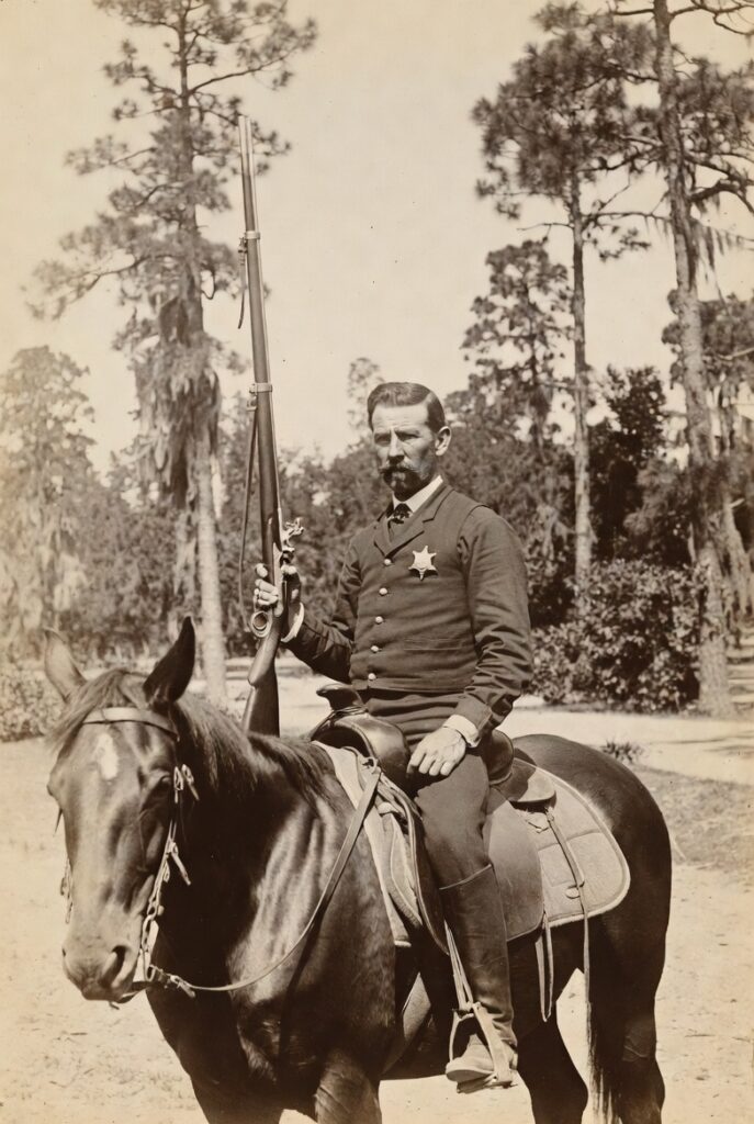 A historical image of a man in uniform seated on a horse, holding a rifle, with a sheriff's badge visible on his chest. The background features wooded trees.