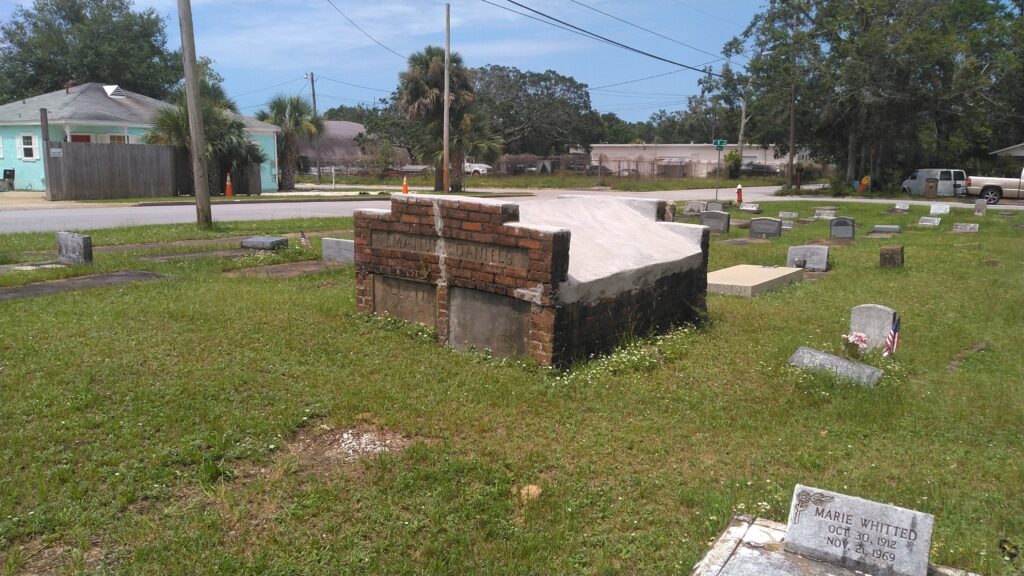 A brick and concrete grave marker in a cemetery, surrounded by grass and smaller gravestones. In the background, there are trees and a low building.