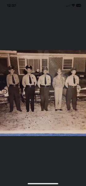 A historical photo from the 1950s showing four law enforcement deputies in uniform standing in front of police cruisers alongside a member of the military.