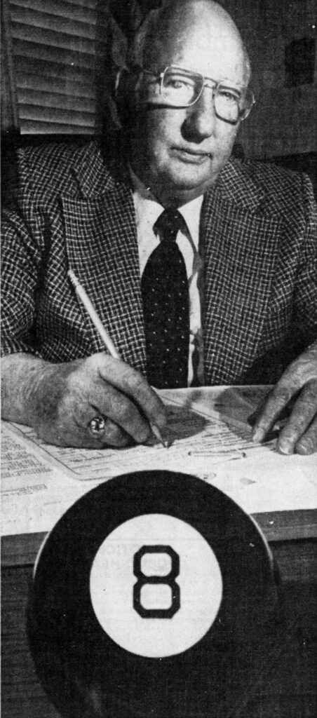 A man with glasses, wearing a checkered suit, sits at a desk writing with a pen. In the foreground, there's an Eight Ball, suggesting a game theme.