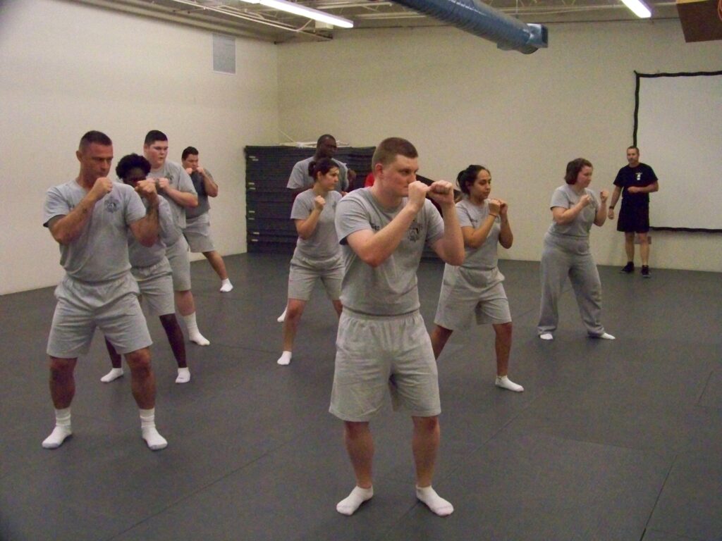 A group of law enforcement academy trainees practicing defensive techniques in a training room, wearing gray uniforms.