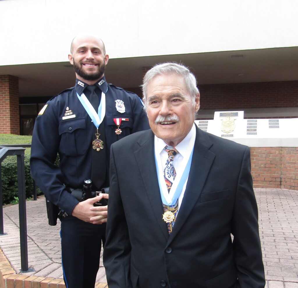 Two men standing outdoors, one in a police uniform smiling and the other in formal attire, both wearing medals around their necks.