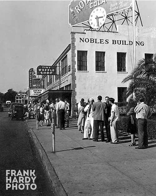 Black and white historical photograph showing a group of people gathered outside the Nobles Building, with signage for Tiny Waffle Shop and other businesses visible.