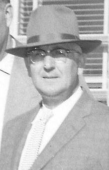 Black and white photo of a middle-aged man wearing a suit and a fedora hat, standing outdoors with a building in the background.