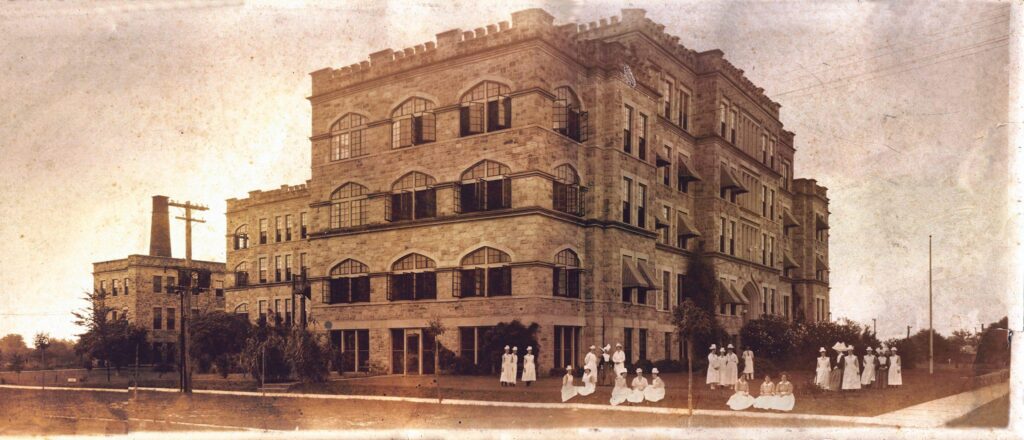 Historic black and white photograph of the Old Pensacola Hospital, featuring a large stone building with multiple windows and an arched entrance, surrounded by trees and several people in white attire gathered on the lawn.