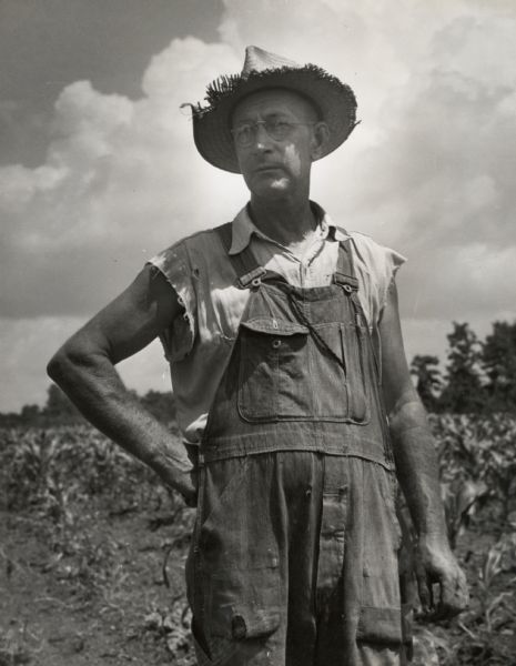 A 58-year-old farmer wearing a straw hat and overalls stands confidently in a field, surrounded by crops and under a partly cloudy sky.
