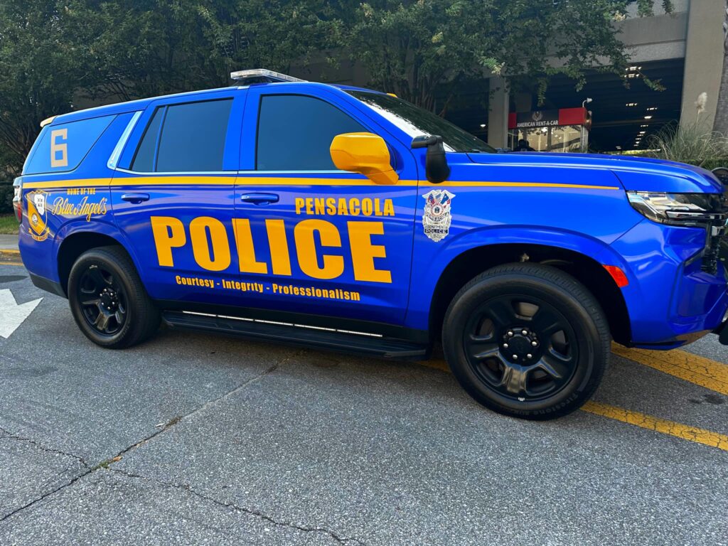 A blue and yellow police vehicle from Pensacola, labeled with 'POLICE' and the Blue Angels insignia, parked on a city street.