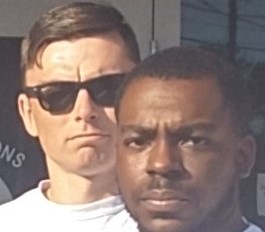Two men standing close together, one wearing sunglasses and a white shirt, and the other with a serious expression.