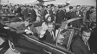 A historical black and white photograph showing John F. Kennedy and Jacqueline Kennedy in a convertible vehicle, waving to a crowd as they arrive at an airport, surrounded by onlookers and airport staff.