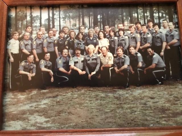 Group photo of law enforcement training class at Pensacola Junior College in the late 1970s, featuring officers in gray uniforms posing outdoors among trees.
