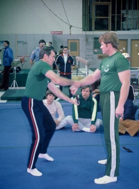 Two male instructors in green shirts demonstrate a training technique on a blue mat, while several students observe in the background.