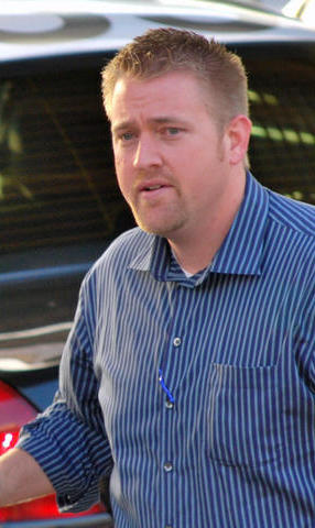 A man with short hair and wearing a blue striped shirt stands in front of a car, appearing thoughtful or engaged in conversation.