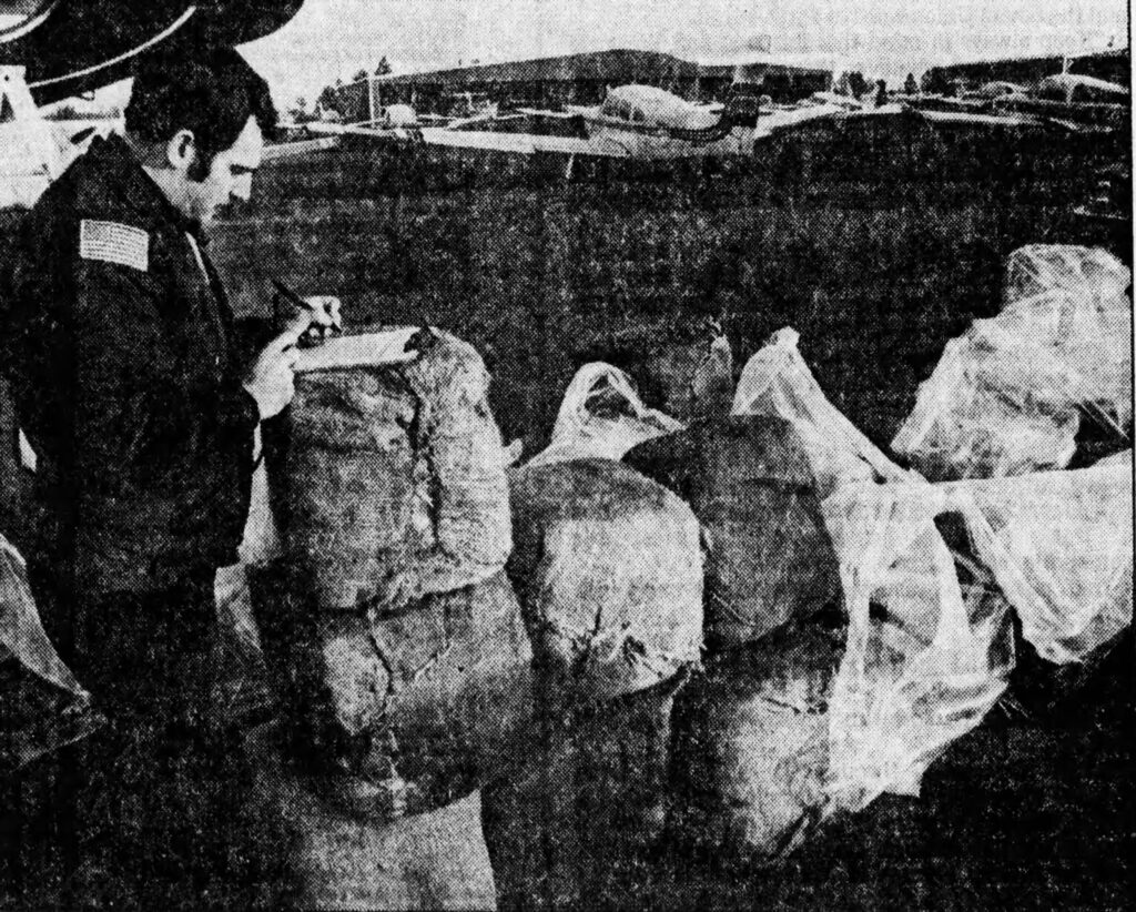 A police officer takes notes next to wrapped bundles of marijuana at an airport, with small planes in the background.