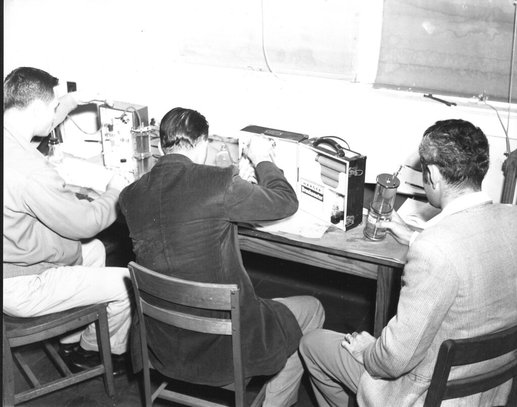 Three men seated at a table, working on early breathalyzer testing equipment.