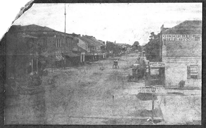 Historical photograph of downtown Pensacola in 1900, showing storefronts, unpaved streets, and early automobiles.
