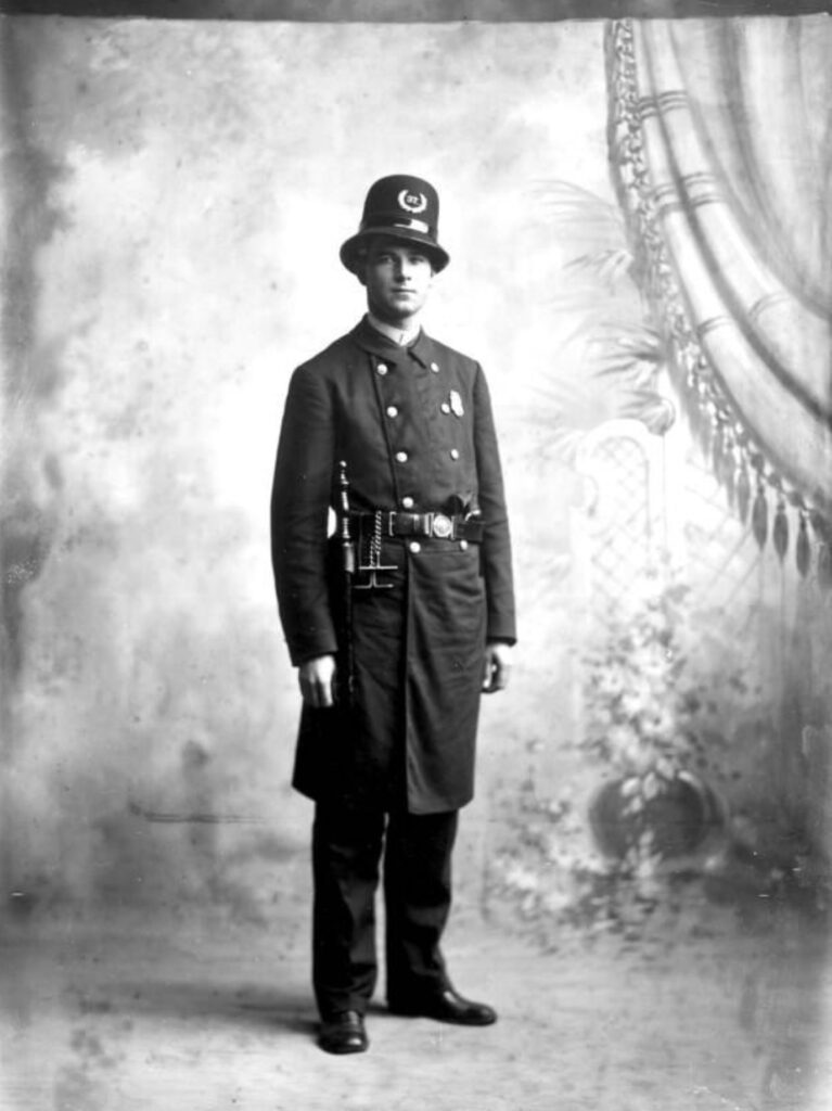 Historical portrait of a male police officer in uniform, standing against a decorative backdrop.