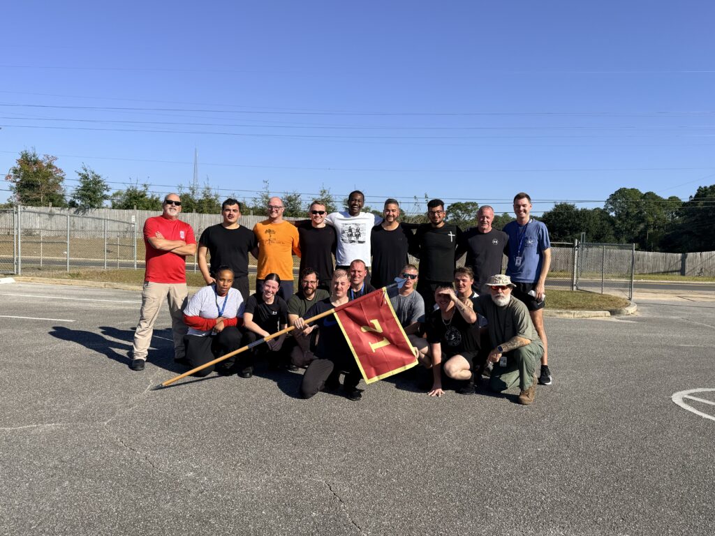 Group photo of a diverse team of individuals posing outdoors, smiling, with a red and yellow flag in front of them. The background features a clear blue sky and a fenced area.