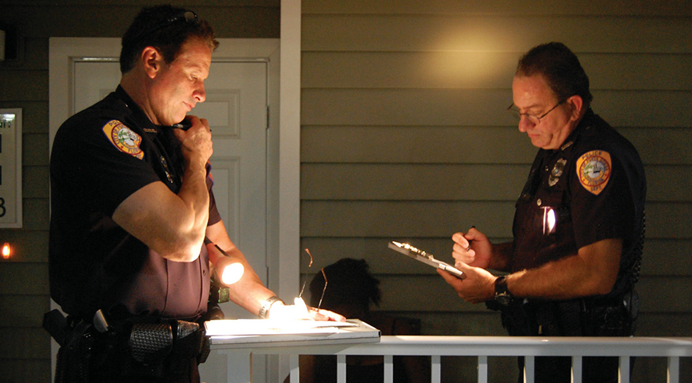 Two police officers discussing on a porch at night, one using a flashlight and the other taking notes.