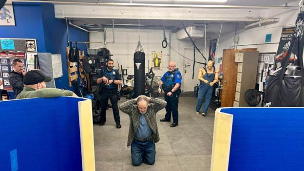 A man kneeling in a gym with his hands behind his head, surrounded by police officers and onlookers. Boxing equipment is visible in the background.