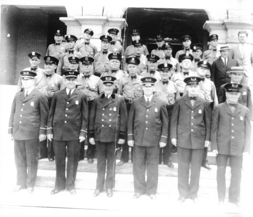 A historic black and white photograph of a group of police officers and municipal workers, posing on the steps of a building. The front row features uniformed officers in formal attire, while the back rows include additional officers in various uniforms.