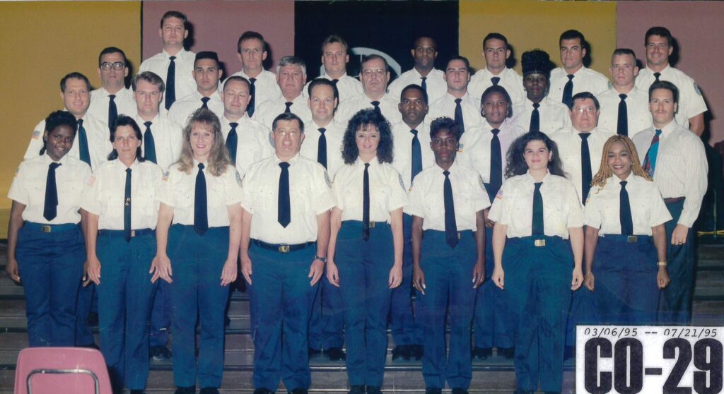 Group photo of an academy class, featuring diverse individuals in uniforms with ties, standing in multiple rows against a colorful background.