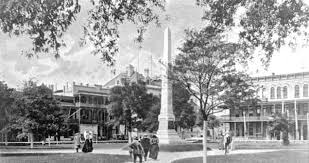 Historic black and white photograph of a park square featuring an obelisk monument, surrounded by trees and early 20th-century buildings, with people walking nearby.