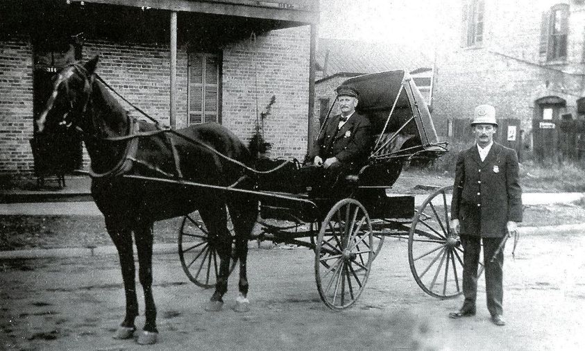 A vintage black and white photograph of a horse-drawn carriage with a seated driver and a man in a uniform standing beside it, with a brick building in the background.