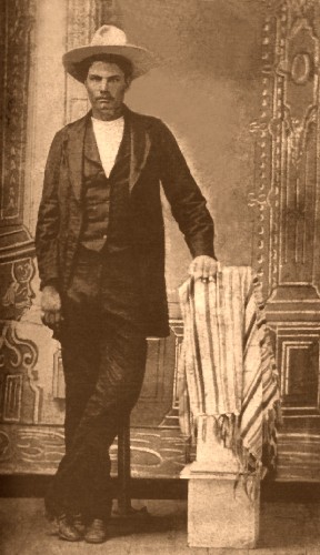 A vintage photograph of a man in a wide-brimmed hat, wearing a dark suit and white shirt, standing beside a draped object on a pedestal.