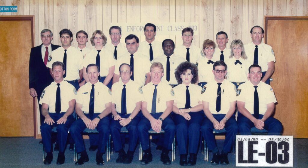 Group photo of law enforcement class LE-03, consisting of male and female trainees in uniforms, posed in front of a wooden wall with a sign indicating 'ENFORCEMENT CLASS #03'. The photo also includes a date range from January 8, 1990, to May 30, 1990.