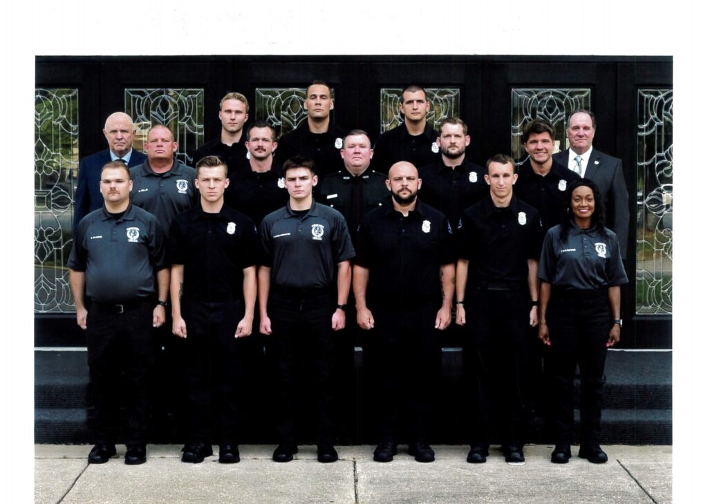 A group photo of law enforcement officers standing in two rows, wearing black uniforms, with a decorative glass door in the background.