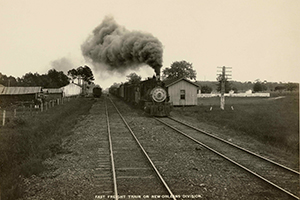 A vintage black and white photograph of a steam locomotive traveling on a railway, with smoke billowing from the engine, and buildings visible alongside the tracks.