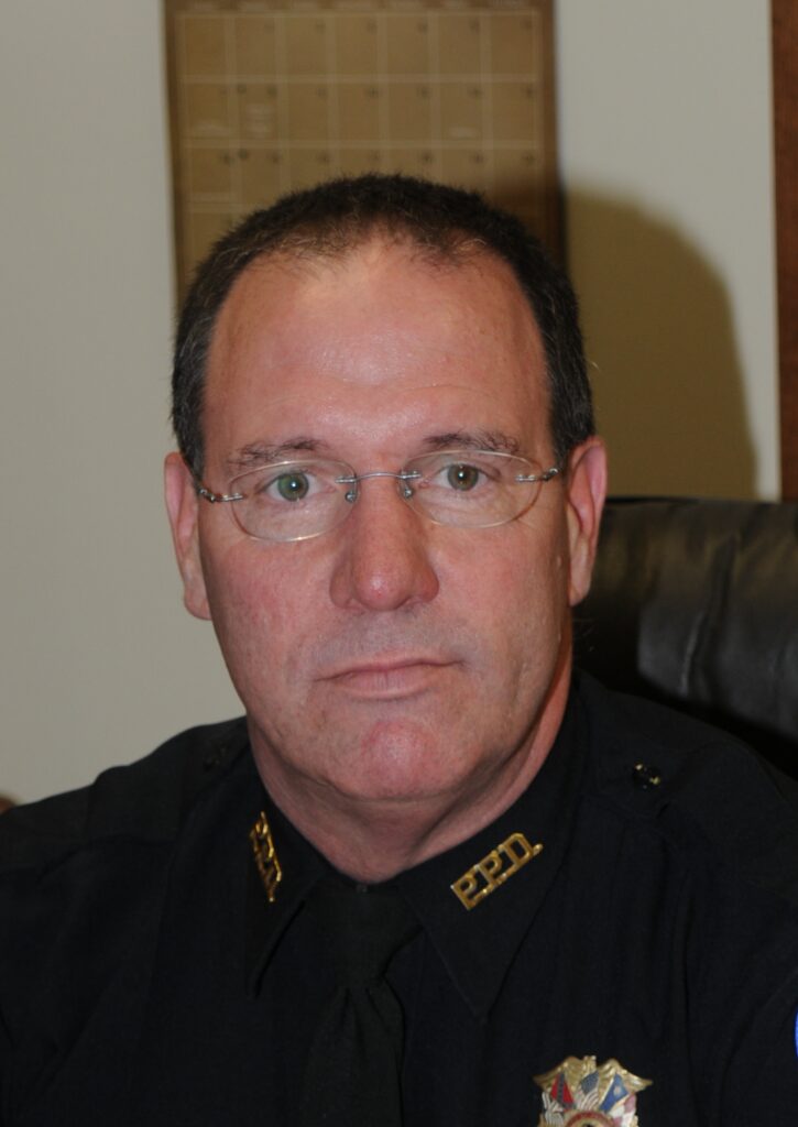 Portrait of a uniformed officer seated at a desk, wearing glasses and a black shirt with a badge.