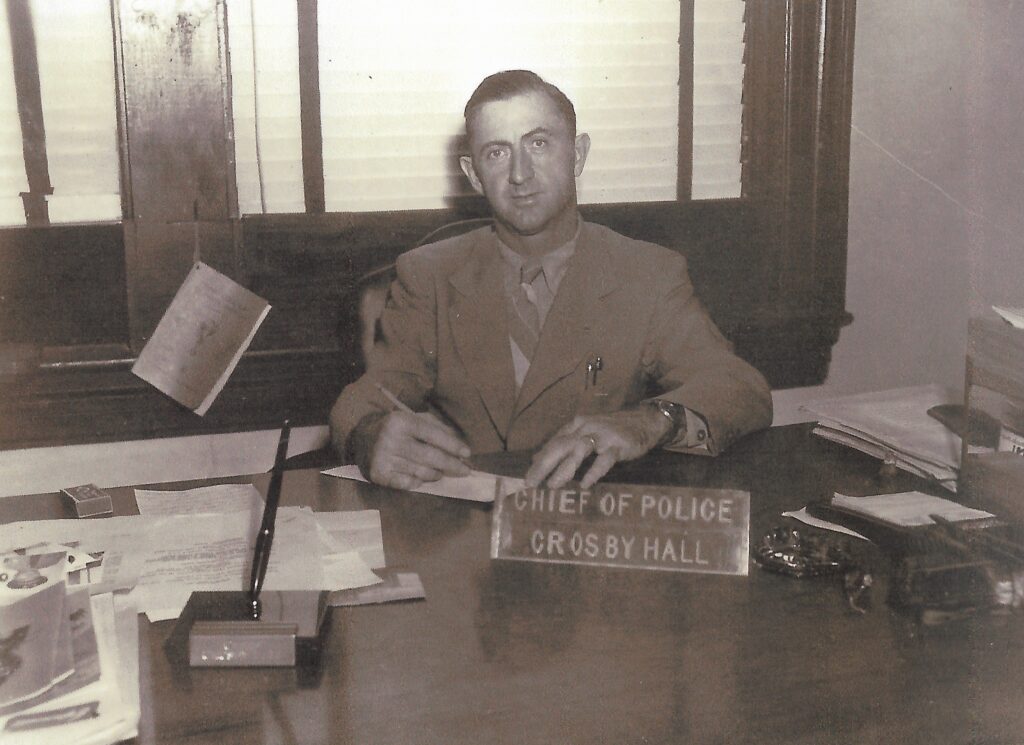 A man seated at a desk in an office, wearing a suit and tie, with a nameplate reading 'Chief of Police Crosby Hall' in front of him.