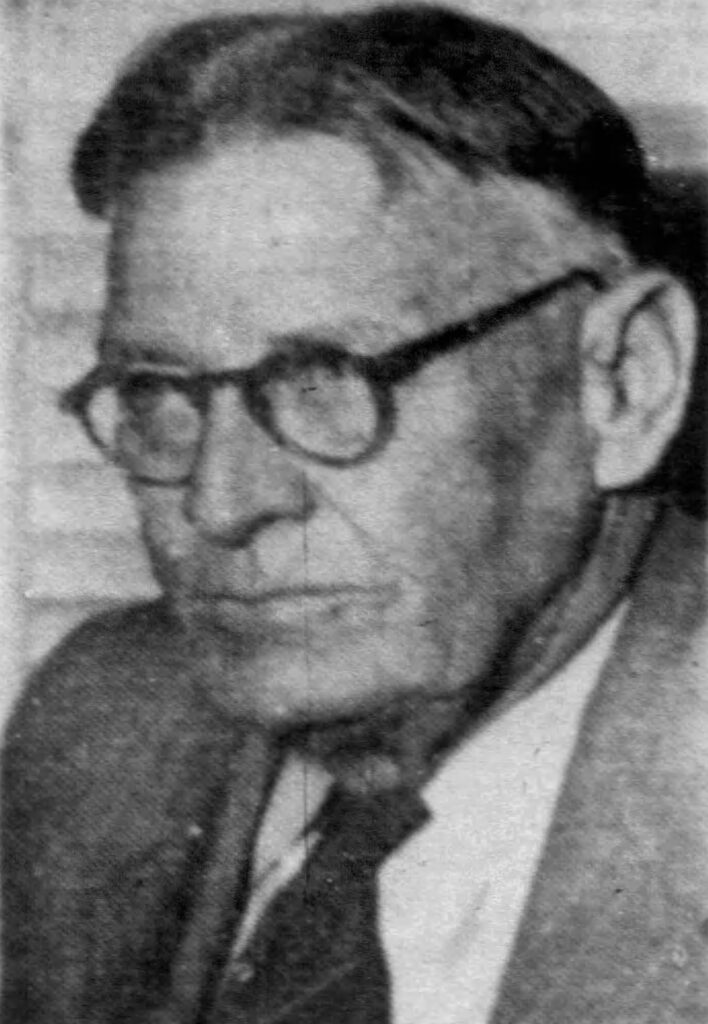 A close-up portrait of an elderly man with glasses, wearing a suit and tie, looking thoughtfully to the side.
