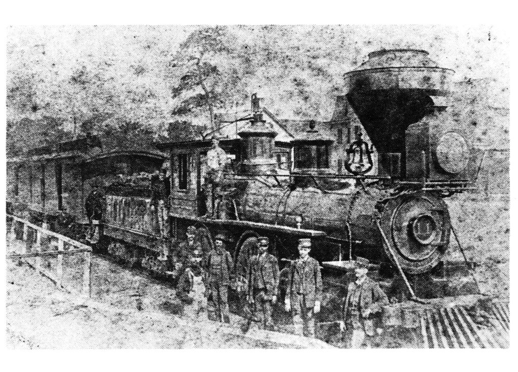 A historical black and white photograph of a steam locomotive with several men in uniform standing beside it. The train is positioned on a track with trees and buildings in the background.
