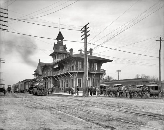 Historic black and white image of a railway station featuring a steam locomotive, a multi-level wooden building with a tower, and horse-drawn carriages parked outside.