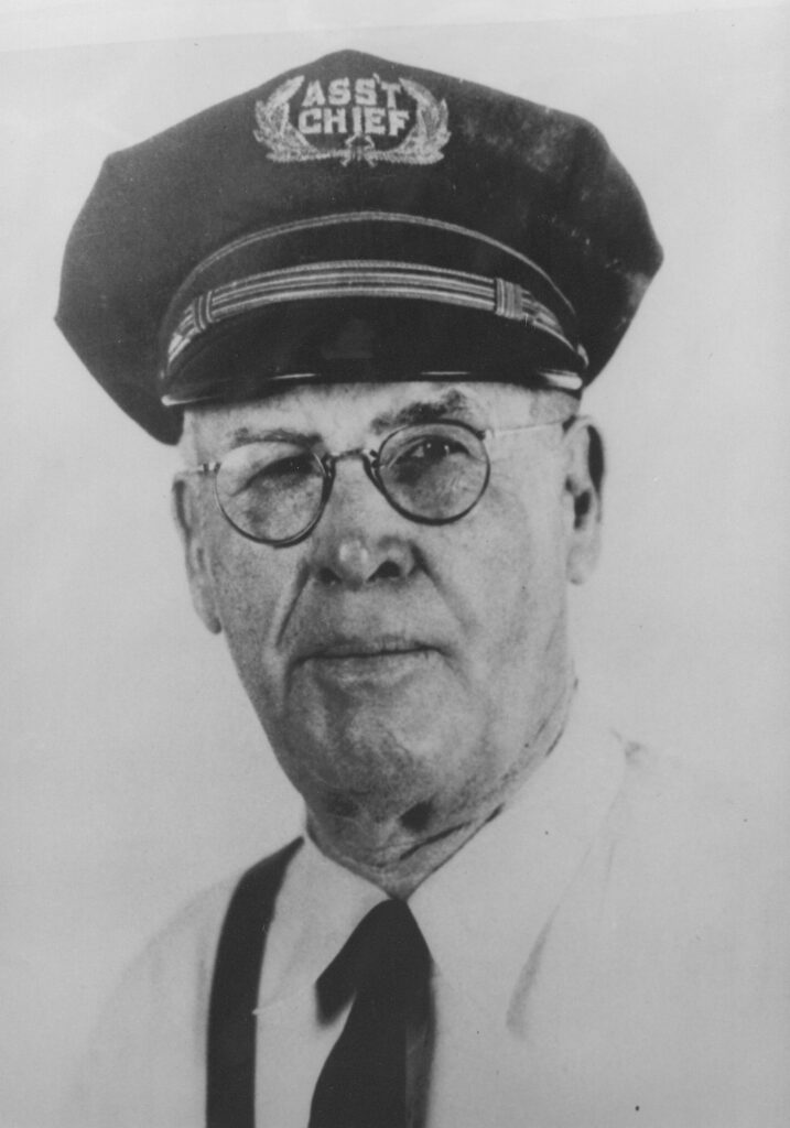 Black and white portrait of an elderly man wearing an 'Assistant Chief' cap and glasses, dressed in a collared shirt with a tie.