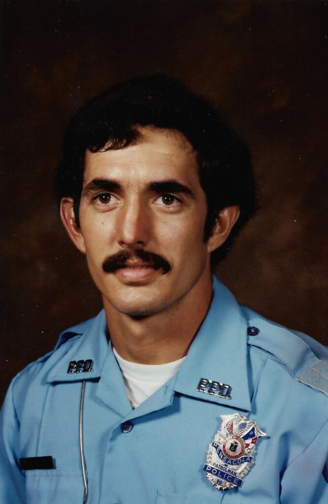 Portrait of a male police officer in a blue uniform with a badge. He has dark hair and a mustache, looking directly at the camera.