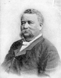 Black and white portrait of a man with a mustache, wearing a suit and bow tie, seated against a plain background.