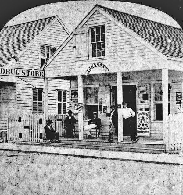 Historic image of a drug store named 'Johnson's,' featuring a group of men gathered outside, with signs visible on the building and a white picket fence in front.