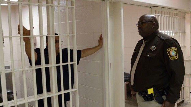 A prison scene with a guard in a brown uniform and a person in a black shirt standing behind bars, reaching out towards the guard.