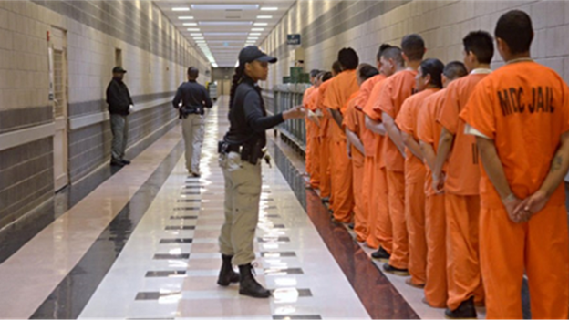 A correctional officer instructs a line of inmates in orange jumpsuits in a prison hallway.