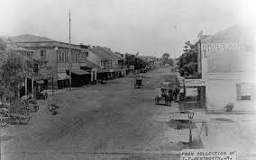 Black and white historical photograph of a street in a small town, featuring buildings on either side, a horse-drawn carriage, and sparse pedestrian traffic.