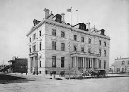 Historic black and white photograph of a large stone building with multiple flags on its rooftop, featuring a columned entrance and horse-drawn carriages in the foreground.