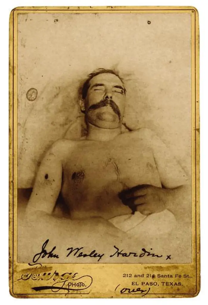 Black and white photograph of a deceased man lying on a bed, with a mustache and eyes closed. The photo features a vintage style with a decorative border and the handwritten inscription 'John Wesley Hardin' at the bottom.