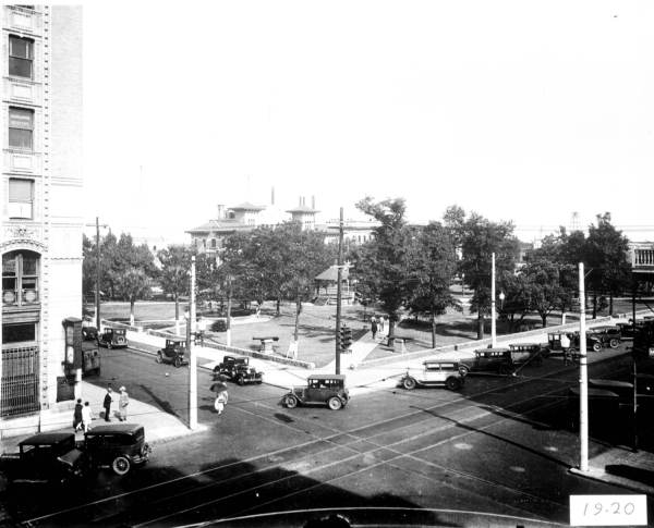 Historic black-and-white photograph of a street intersection in 1920, featuring early model cars and trees in the surrounding park.