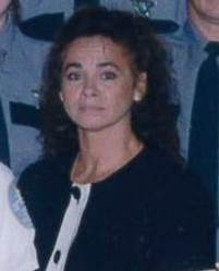 A woman with curly hair wearing a black and white jacket, standing among individuals in uniform, with a serious expression.