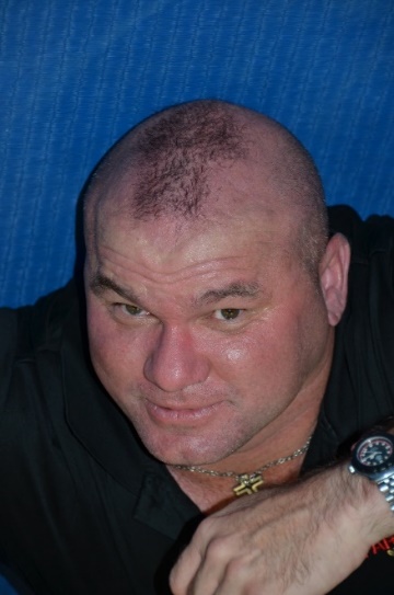 Close-up portrait of a man with a shaved head and short hair on top, wearing a black shirt and a watch, with a serious expression against a blue background.