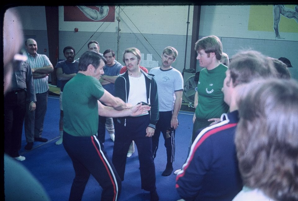 A group of individuals observing a demonstration of a physical technique or martial art in a gymnasium setting, with one person in a green shirt demonstrating while others attentively watch.