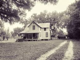 A vintage-style farmhouse with a porch, surrounded by trees and an open yard, captured in black and white.